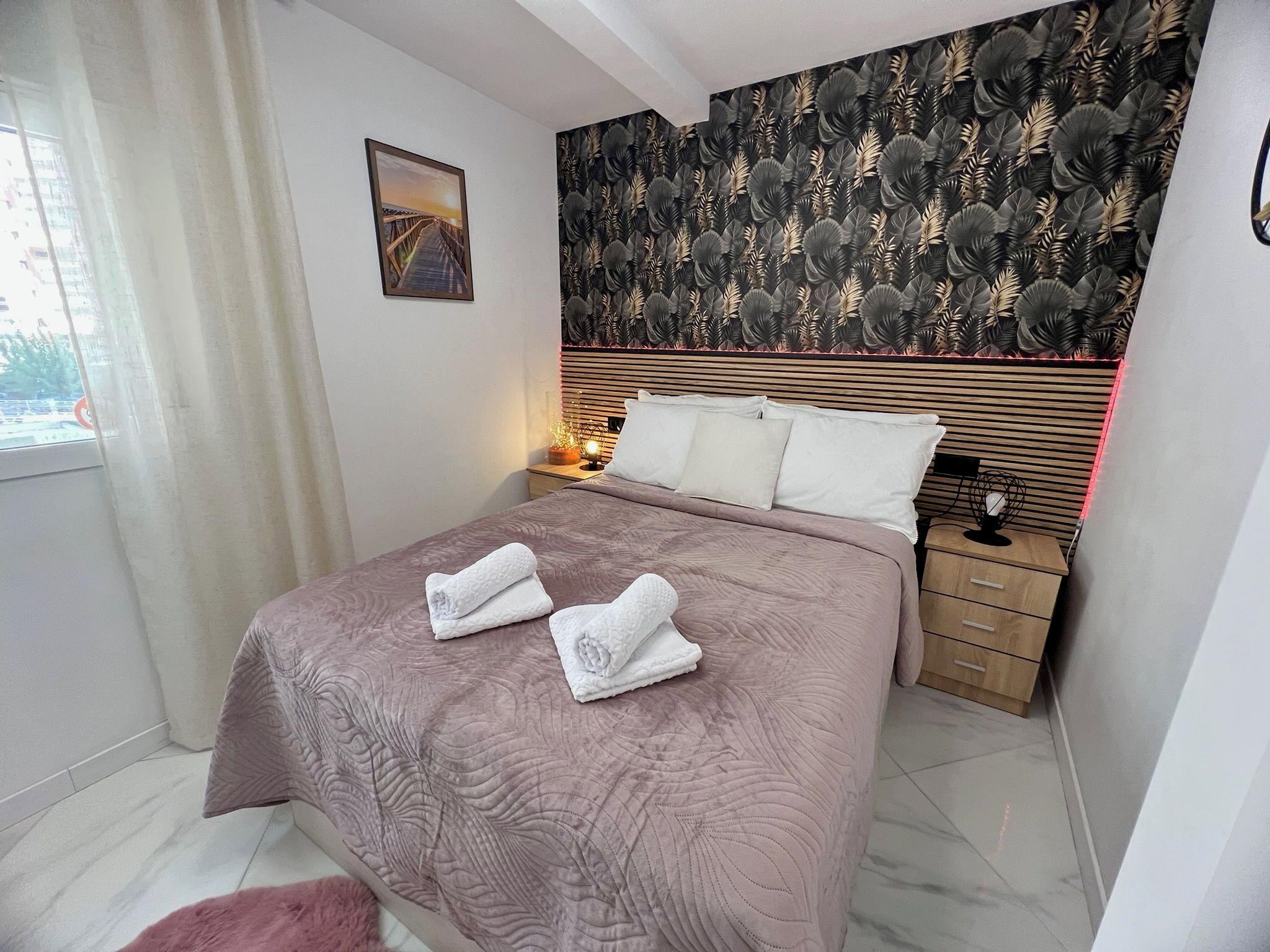 A hotel room with a bed, a nightstand, a lamp, a painting, and a patterned wall.