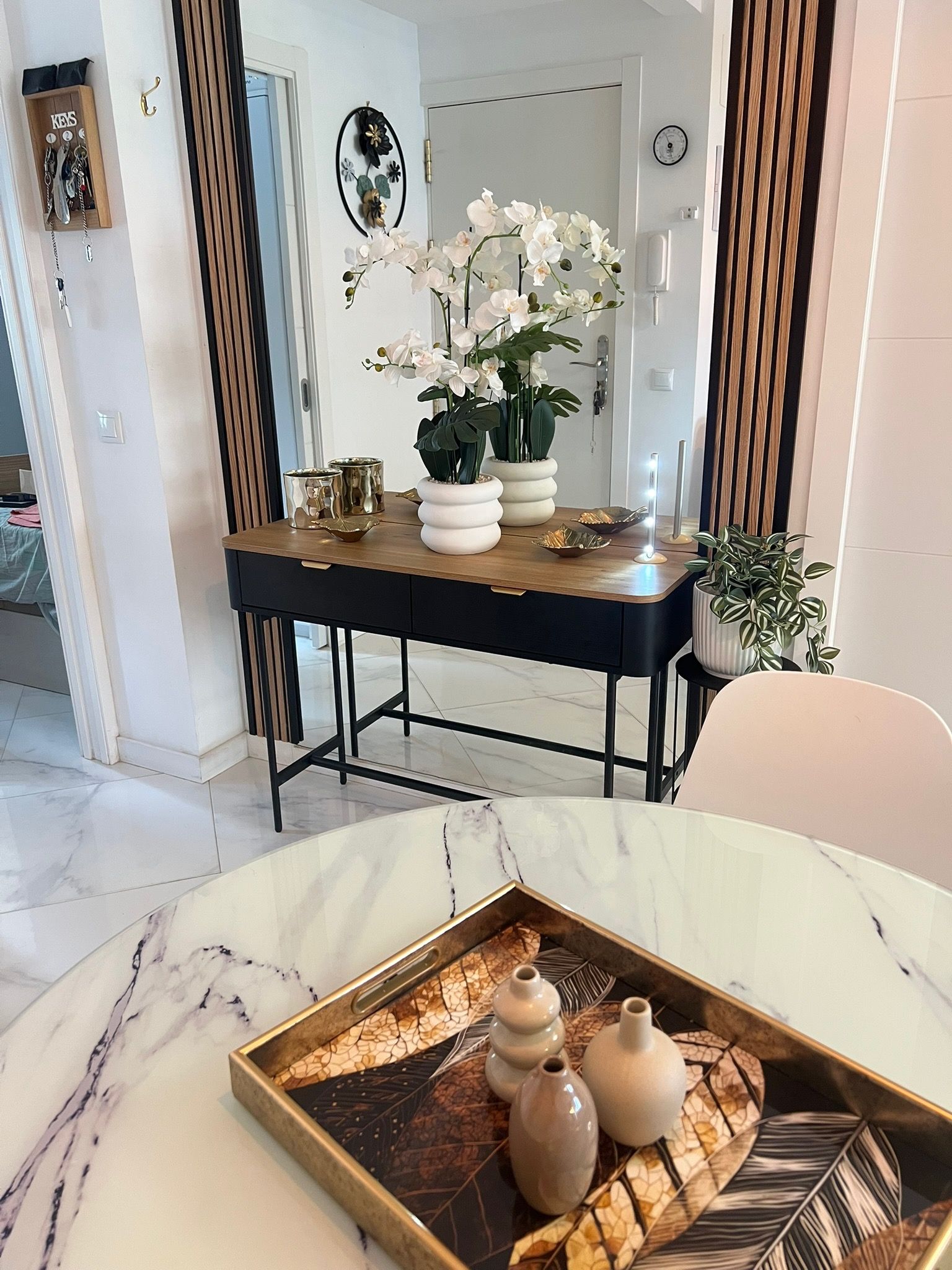 A modern interior with a marble table, a decorative tray with vases, a potted plant, and a wall-mounted key holder.