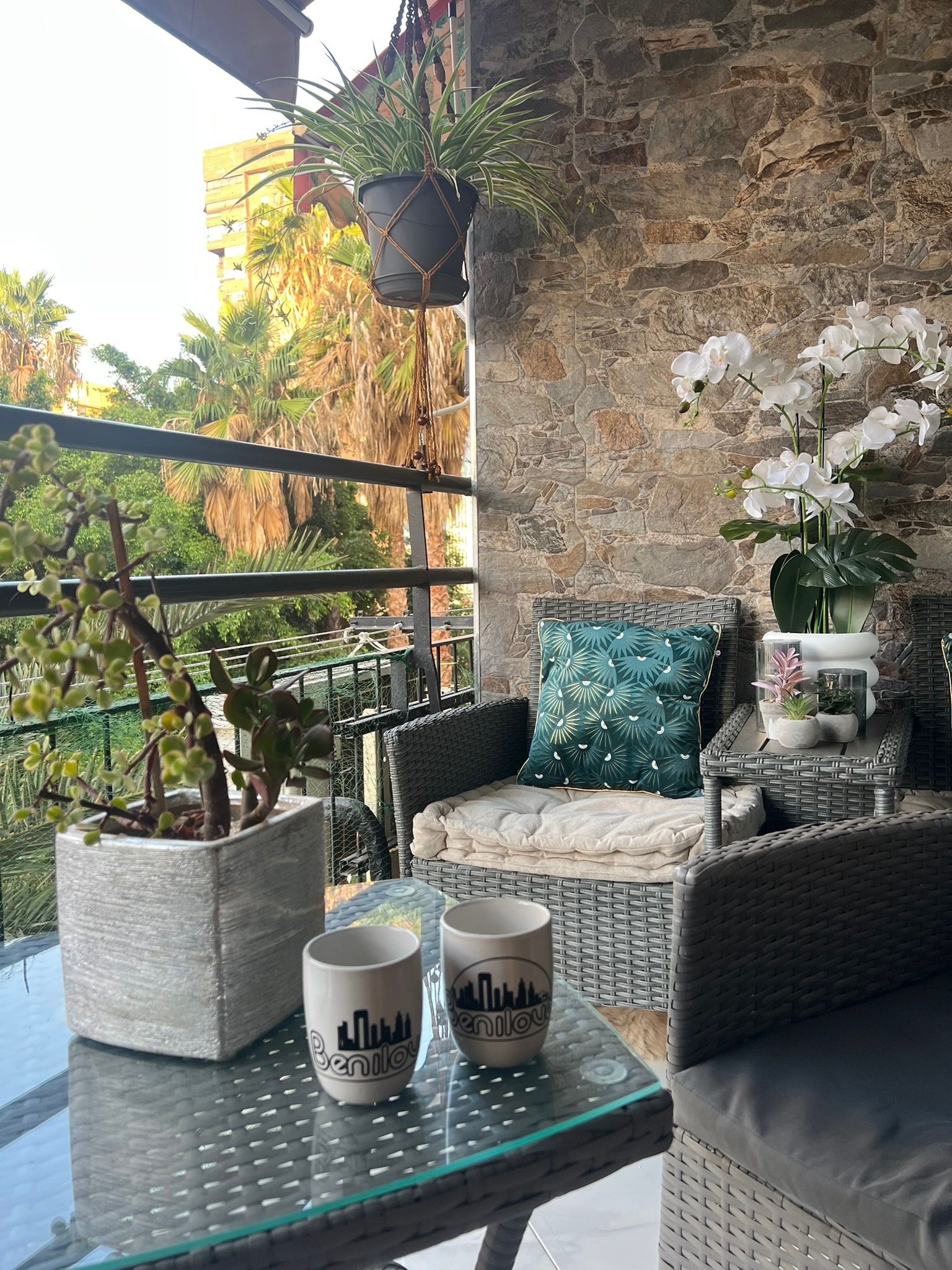 Ein Balkon mit Korbmöbeln, Blumentöpfen und einem Glastisch mit zwei Tassen.