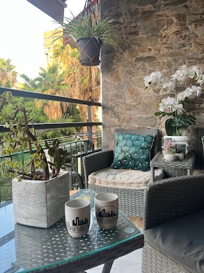 Ein Balkon mit Korbmöbeln, Blumentöpfen und einem Glastisch mit zwei Tassen.