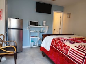 Quarto com cama, frigorífico, micro-ondas, prateleira com utensílios de cozinha e televisão na parede.