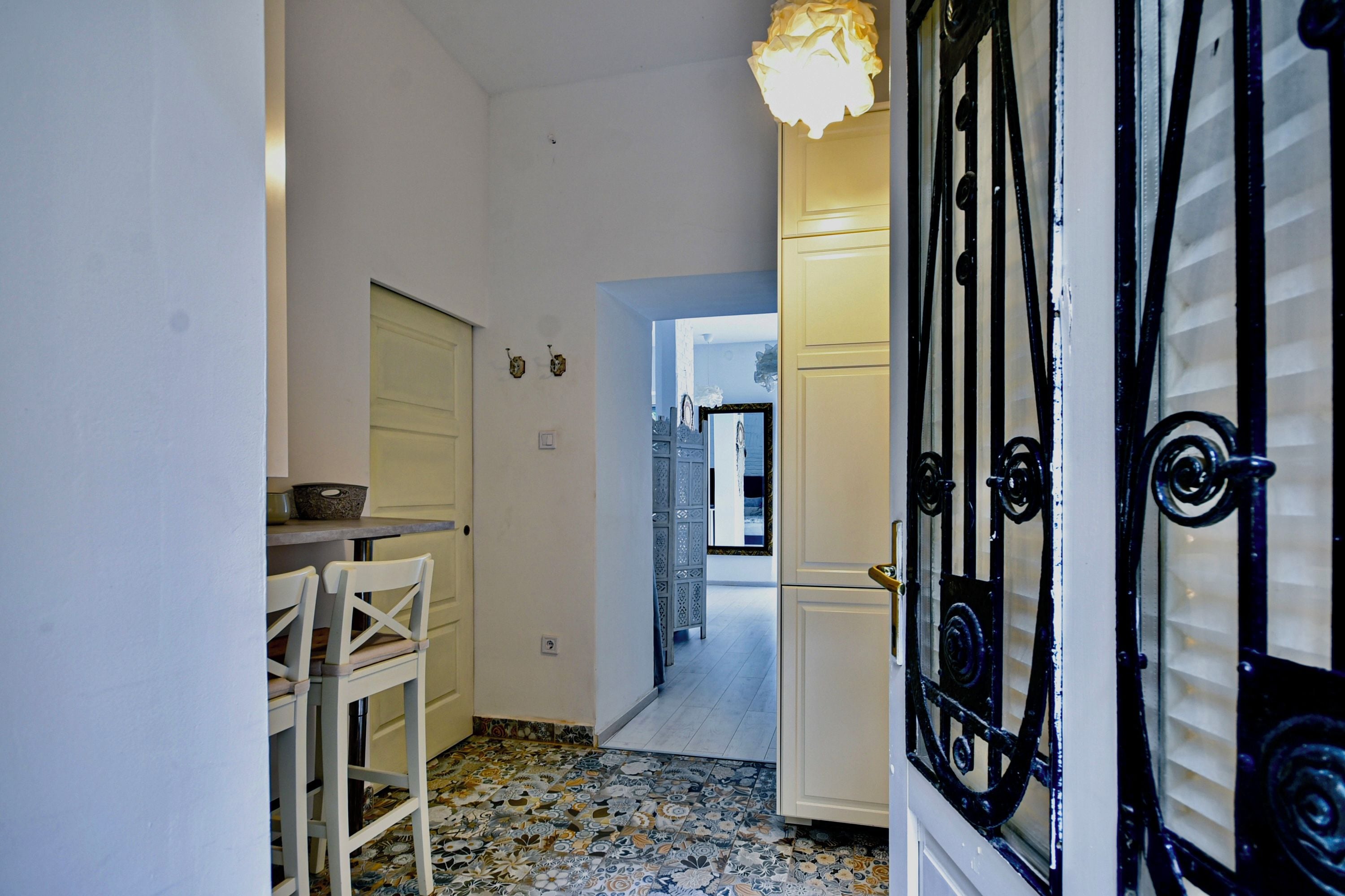 A narrow hallway with a tiled floor, a white door with black metalwork, and a white table with chairs.