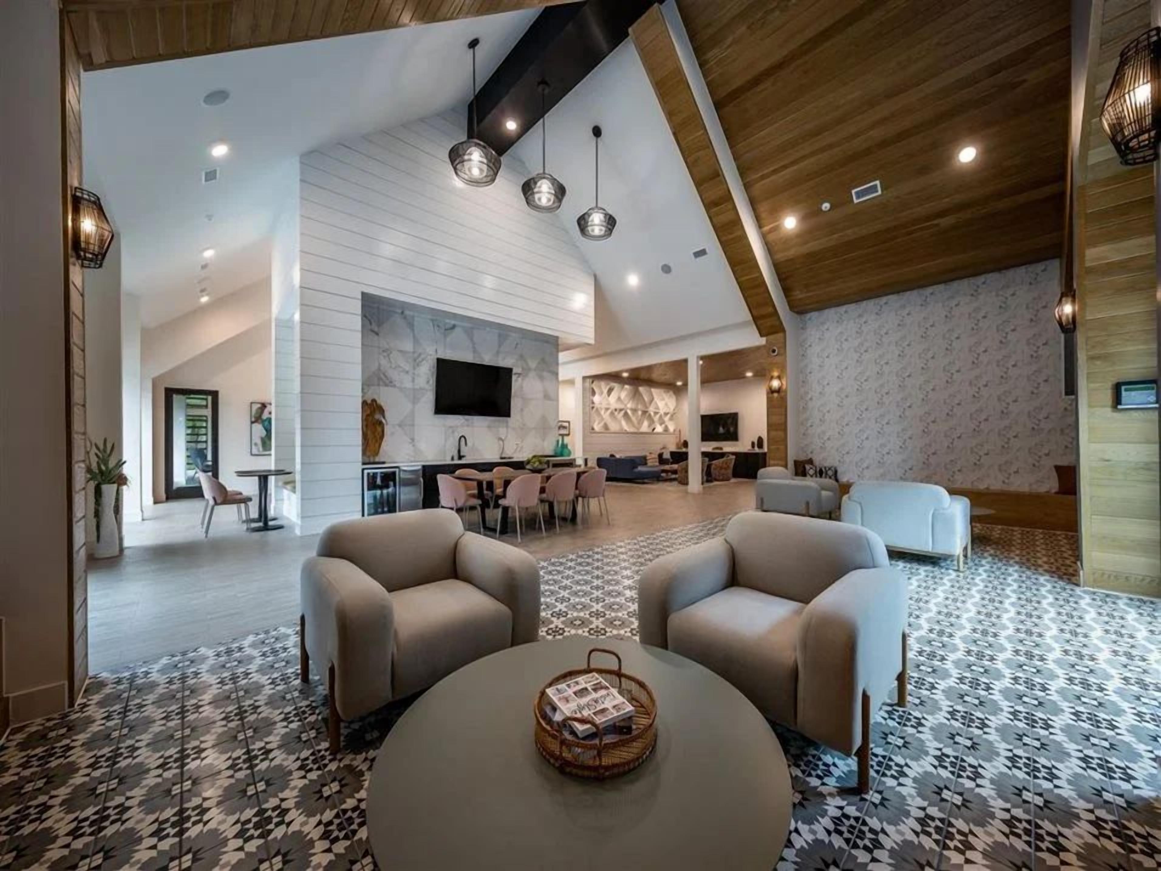 A spacious living area with a modern design, featuring a central seating arrangement, a flat-screen TV mounted on the wall, and a dining area with a kitchen in the background.