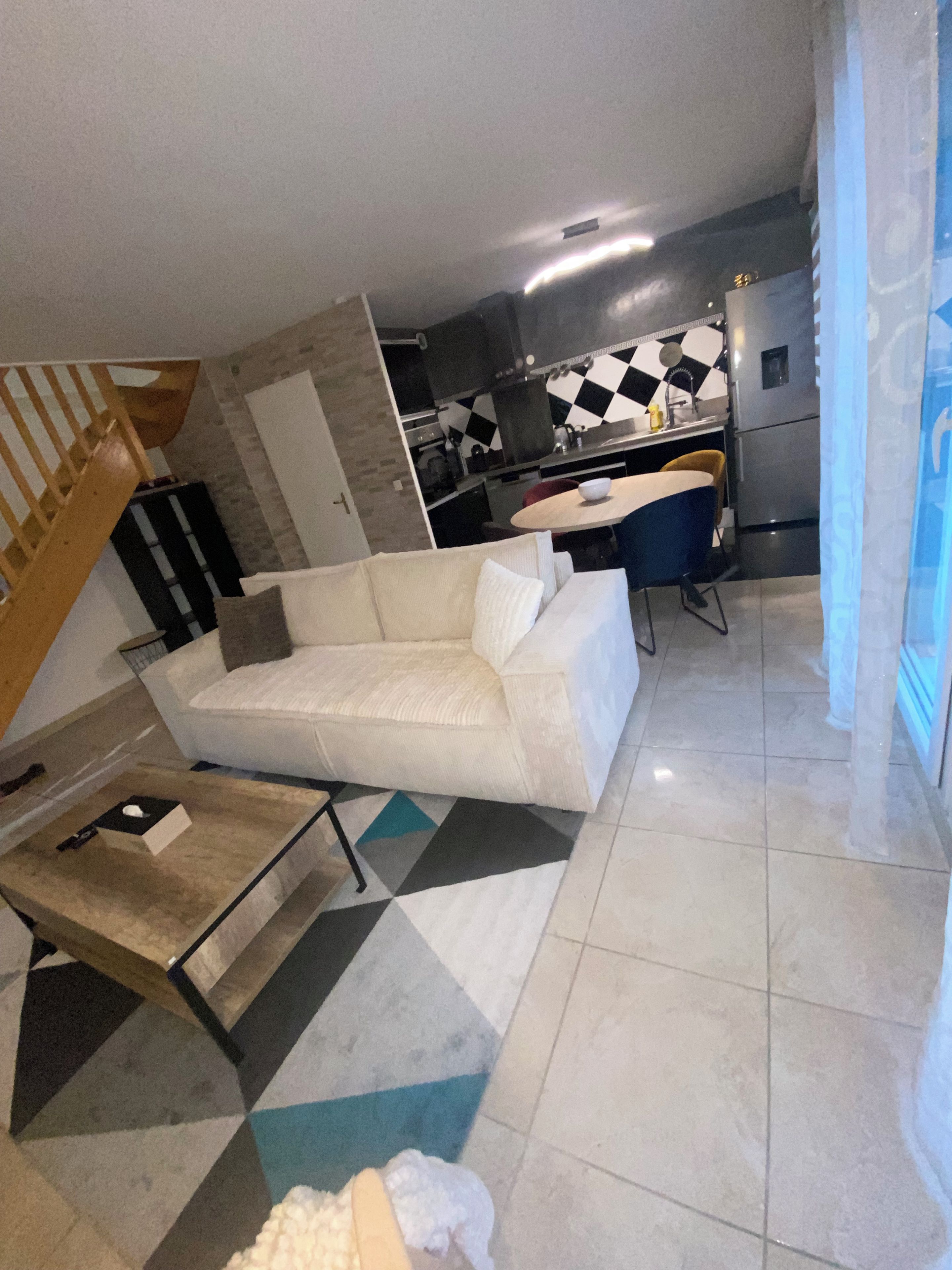A modern living room with a white sofa, a coffee table, and a geometric-patterned rug. A kitchen with black and white checkered backsplash and stainless steel appliances is visible in the background.