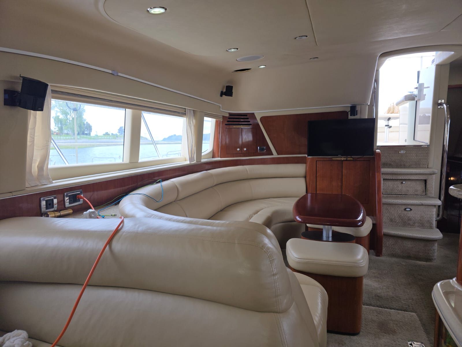 Interior de un barco con una zona de estar curva, un televisor y una escalera que lleva a una cubierta superior.