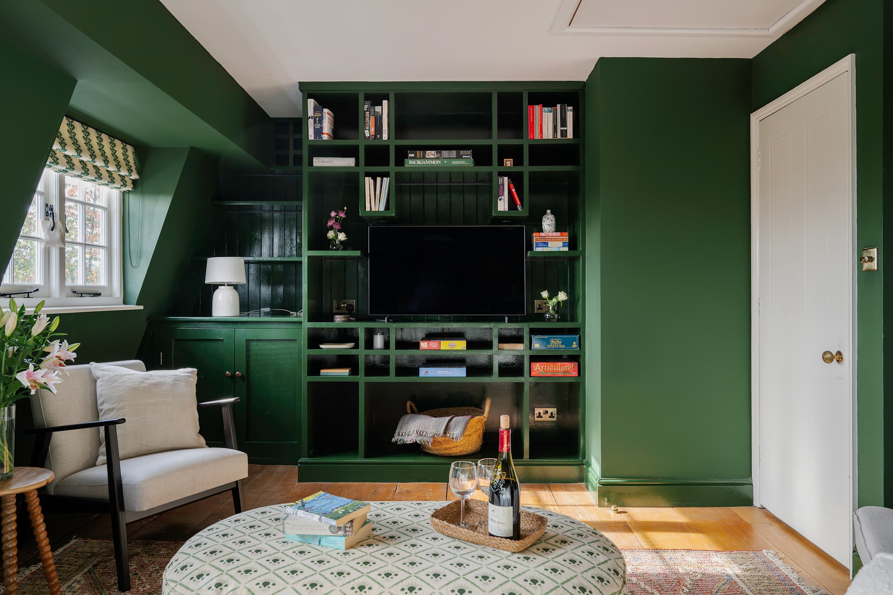 Una sala de estar acogedora con una estantería verde, un sillón cómodo, una mesita con libros y una botella, y un televisor.