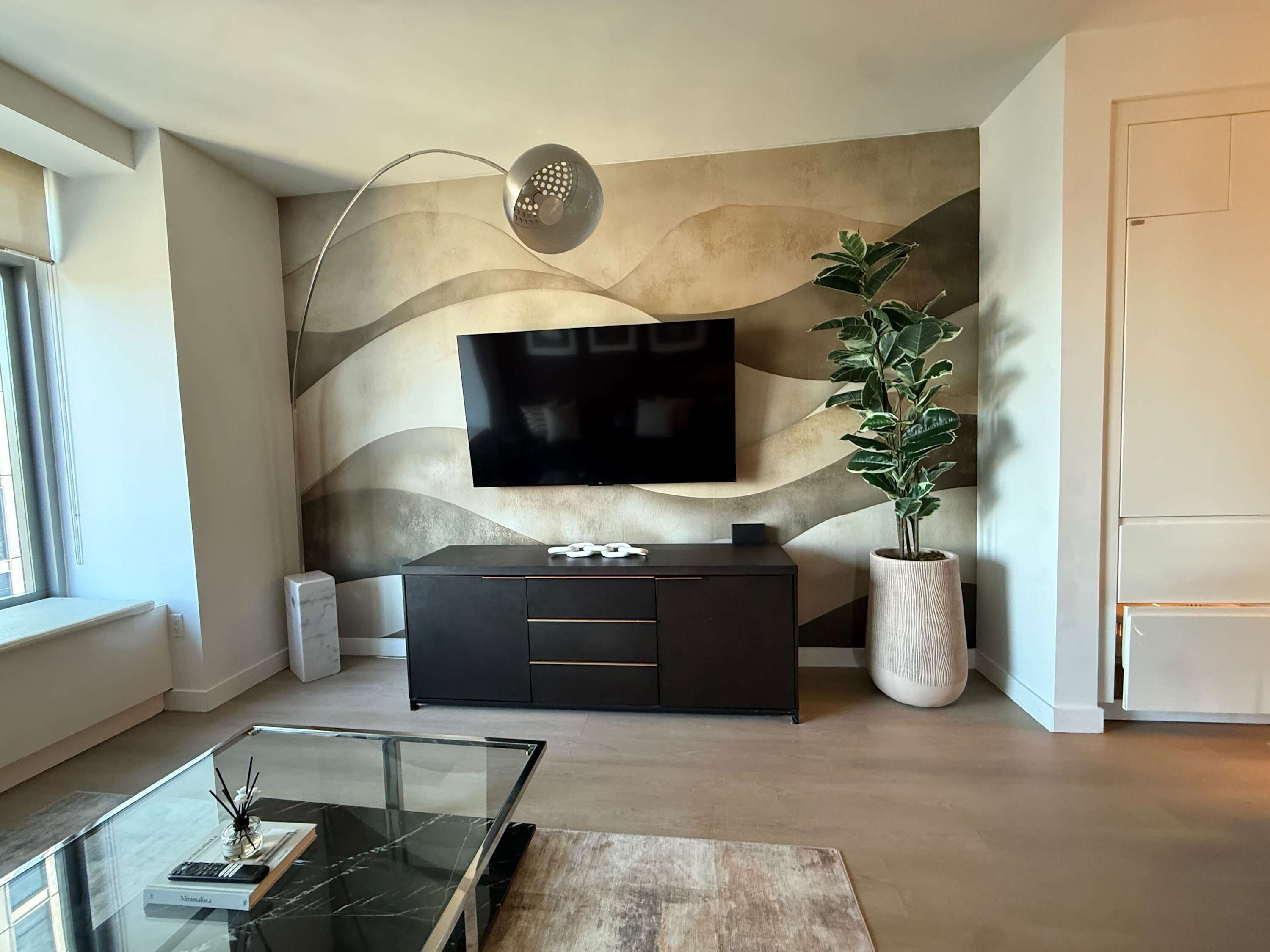 A modern living room with a flat-screen TV mounted on a textured wall, a dark wooden cabinet, a glass coffee table, and a potted plant.