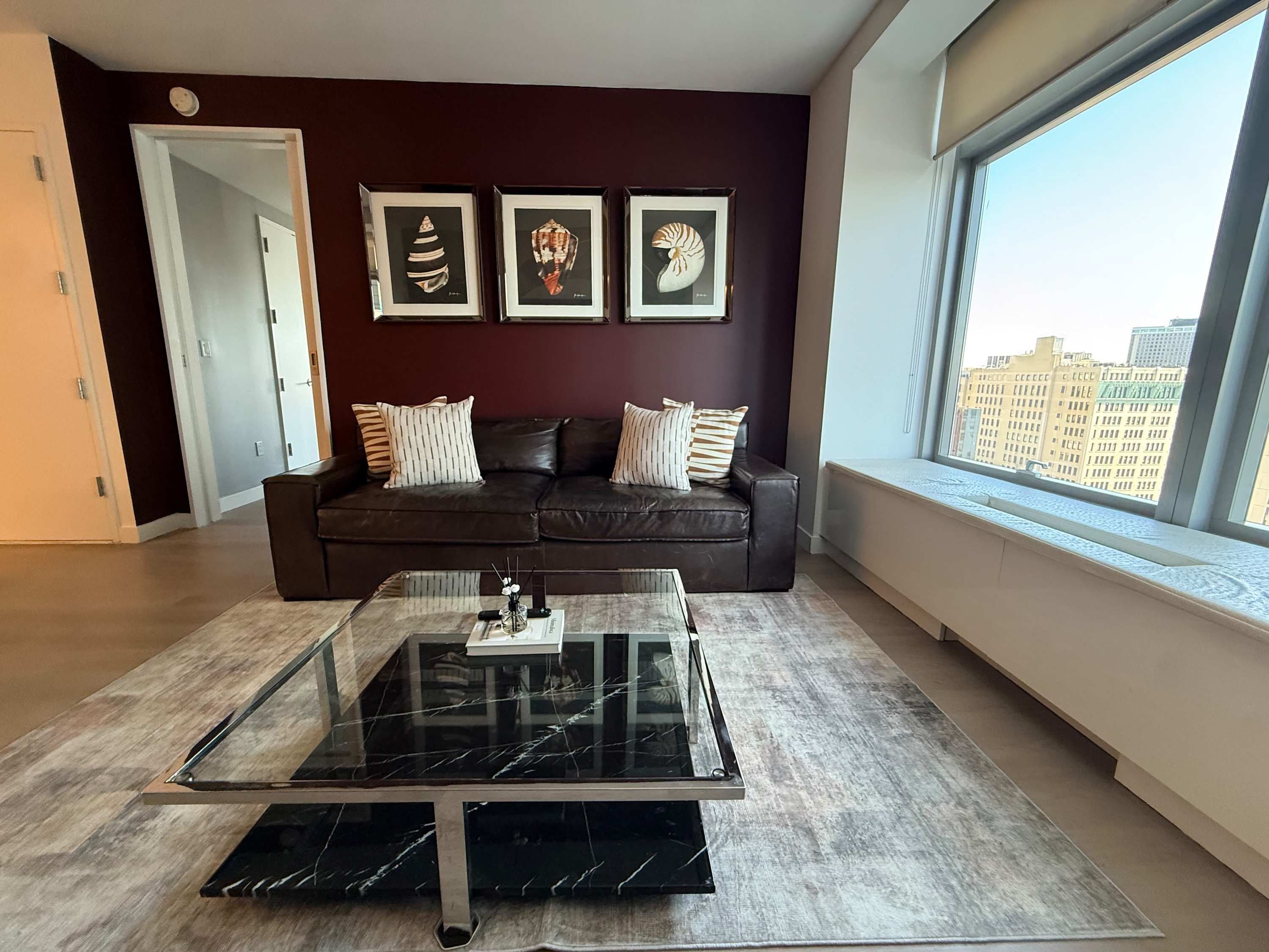 A modern living room with a glass coffee table, a dark brown leather sofa, and framed artwork on the wall.