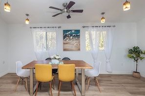 Un comedor con mesa de madera, cuatro sillas, ventilador de techo y dos ventanas con cortinas translúcidas.