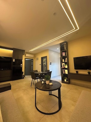 Un salon moderne avec une table basse ronde, un coin repas avec des chaises, une cuisine équipée d’un réfrigérateur et un téléviseur à écran plat fixé au mur.