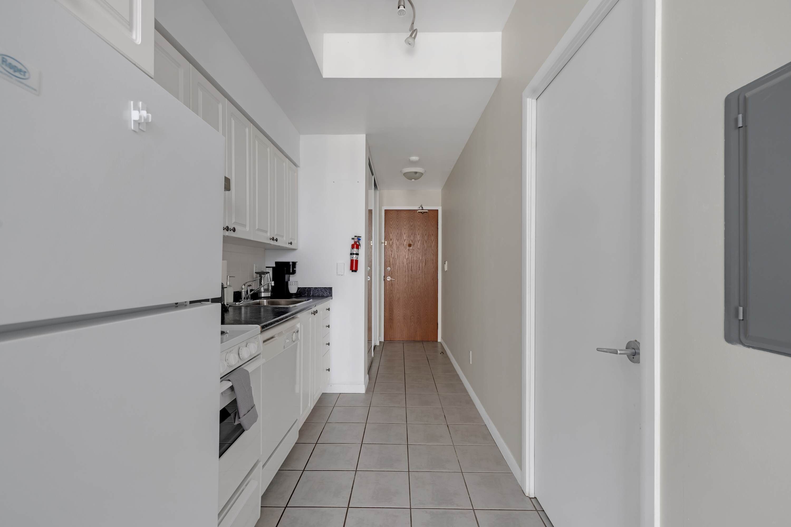 Un couloir étroit, avec une cuisine d’un côté, une porte en bois au fond, et un extincteur fixé au mur.