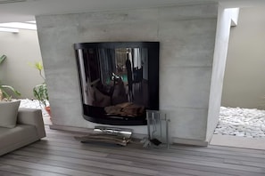 A modern living room with a flat-screen TV mounted on a concrete wall, a sofa, and a fireplace with logs. - Villa Yaya (Hammamet)