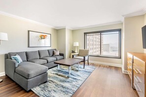 Un salon moderne avec un canapé gris, une table basse, un tapis, un fauteuil, une lampe et une grande fenêtre donnant sur la ville.