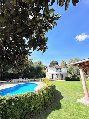 Une piscine entourée d’une pelouse bien entretenue, un bâtiment en pierre avec un toit en tuiles et une structure en forme de pergola.