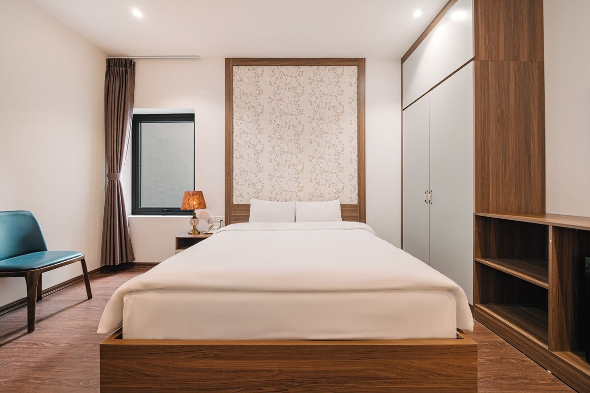 A bedroom with a bed, a nightstand, a lamp, a chair, a wardrobe, and a window with curtains.