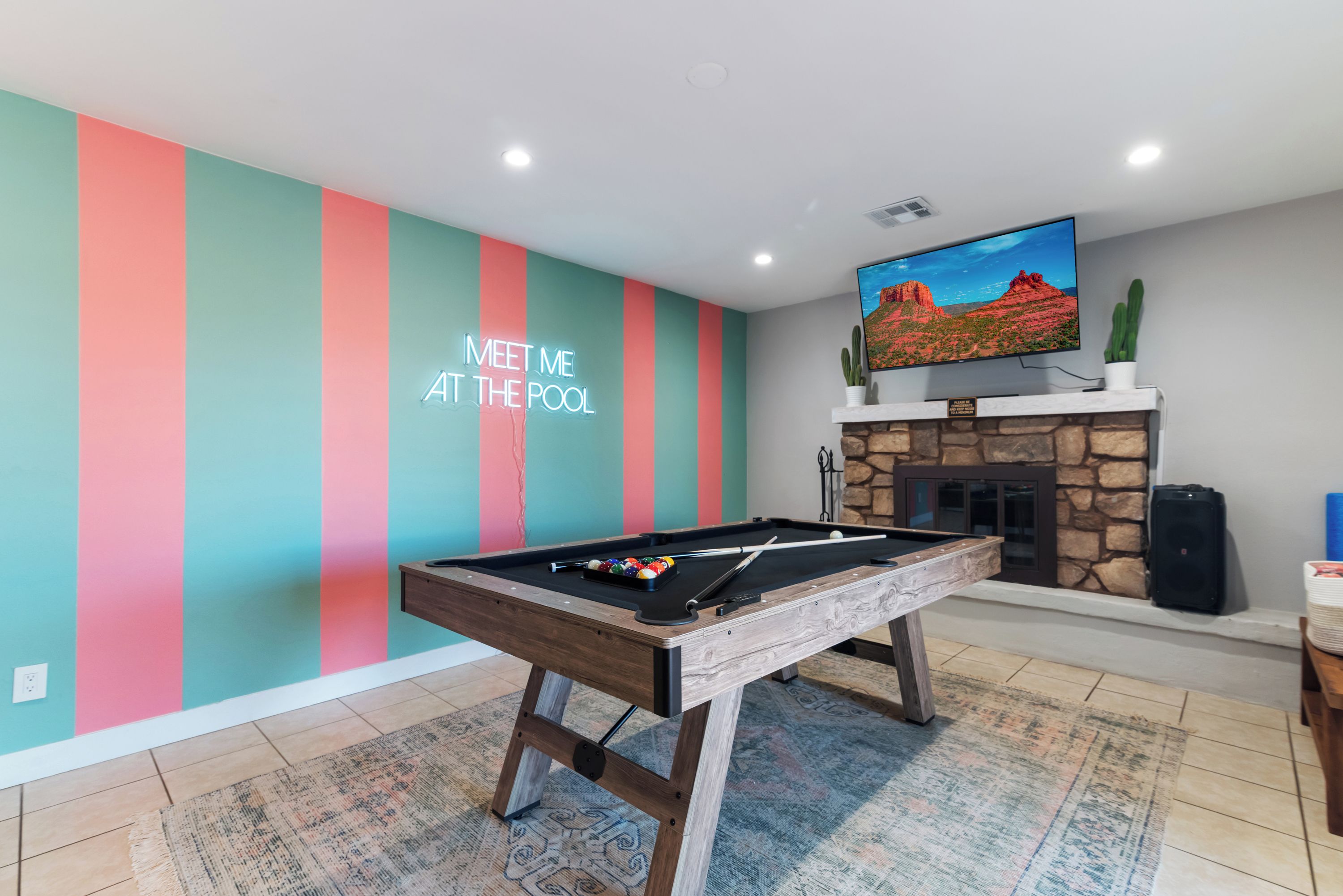 Une table de billard dans une pièce dont un mur est décoré de rayures colorées, et où un téléviseur fixé au mur diffuse des images d’un paysage désertique.