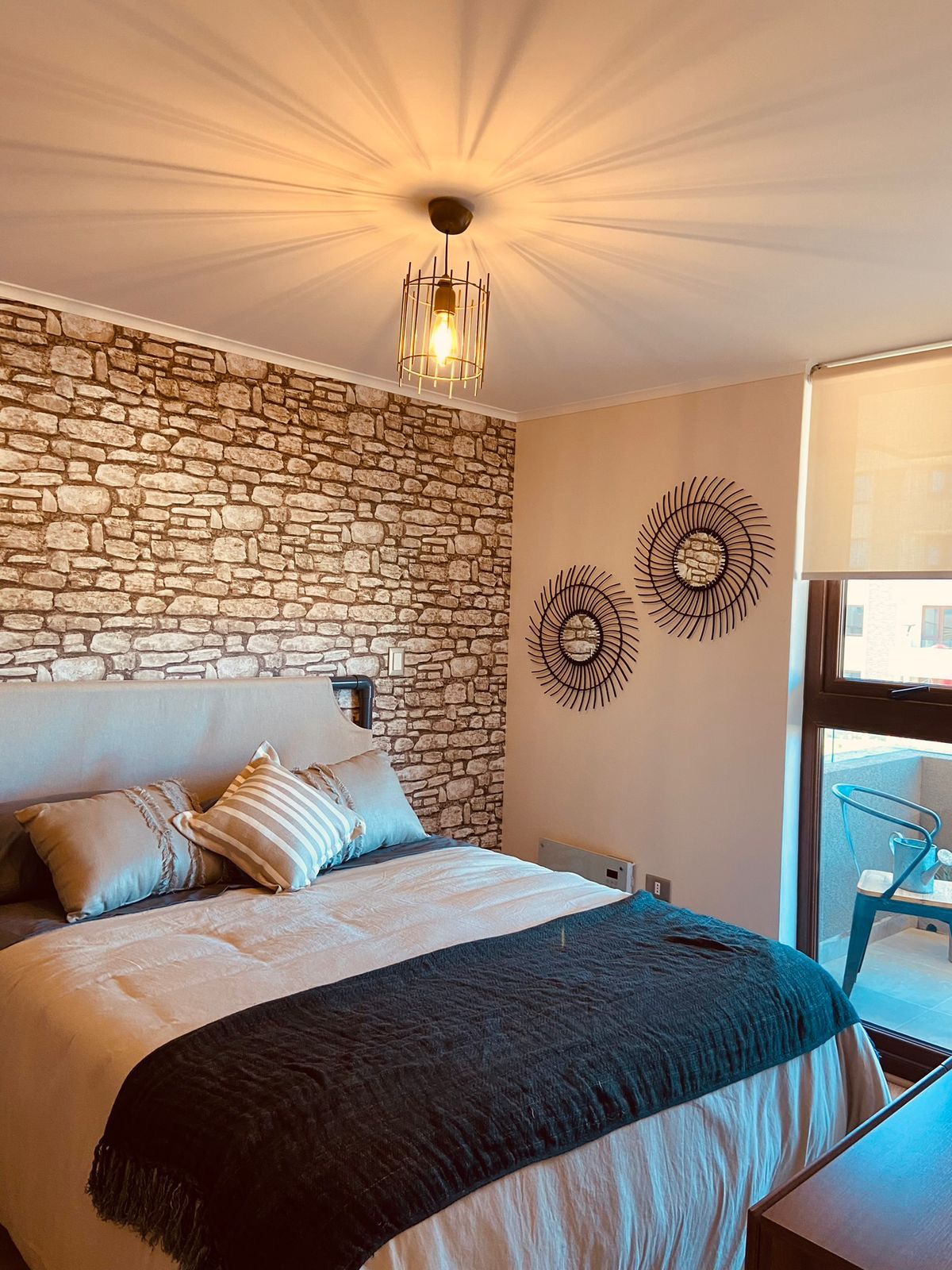 A bedroom with a stone wall, a bed with pillows, a hanging light fixture, and a view of the outside.