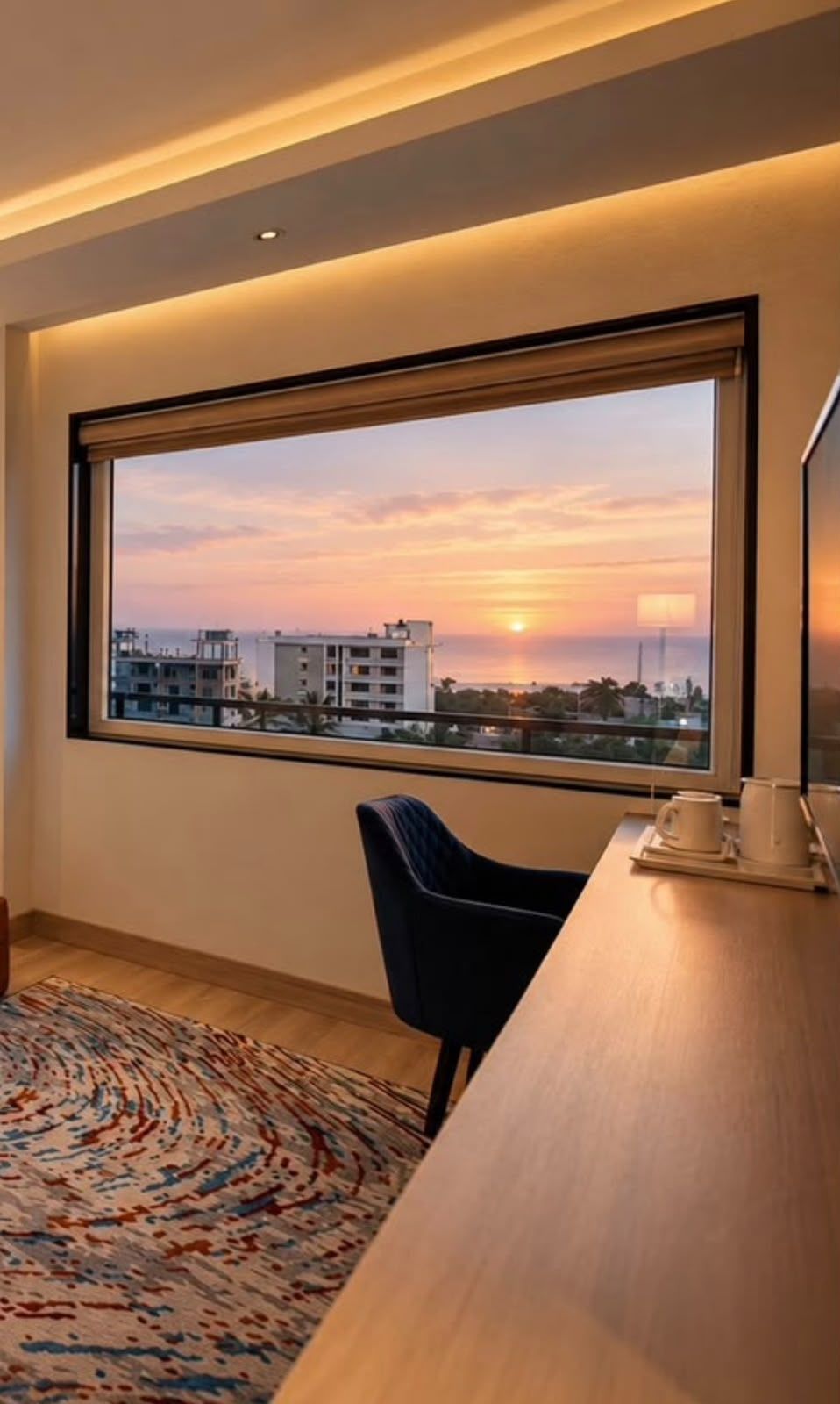 A room with a large window, a wooden desk, a blue chair, and a patterned rug.