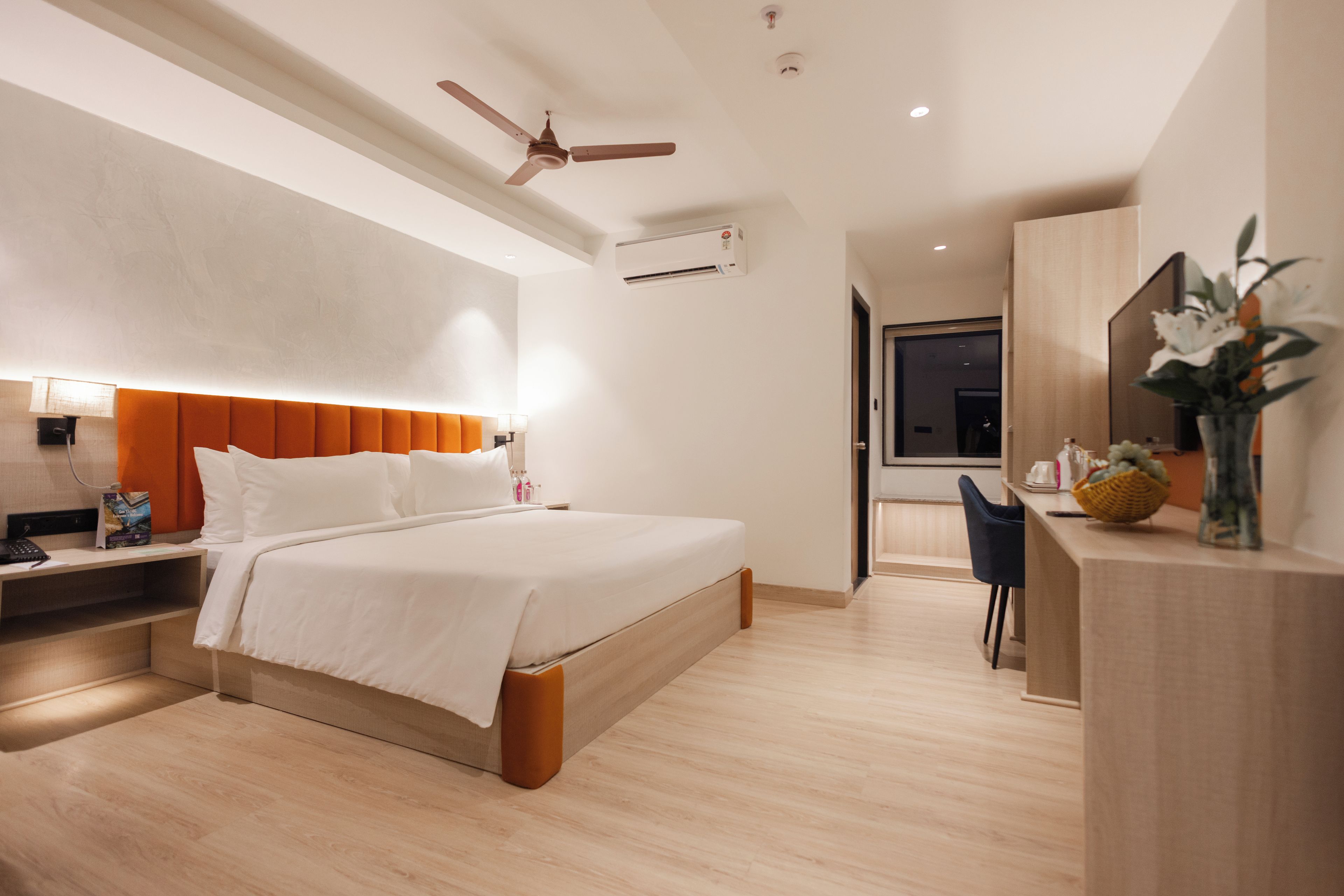 A modern hotel room with a bed, bedside table, lamp, and a desk with a chair.