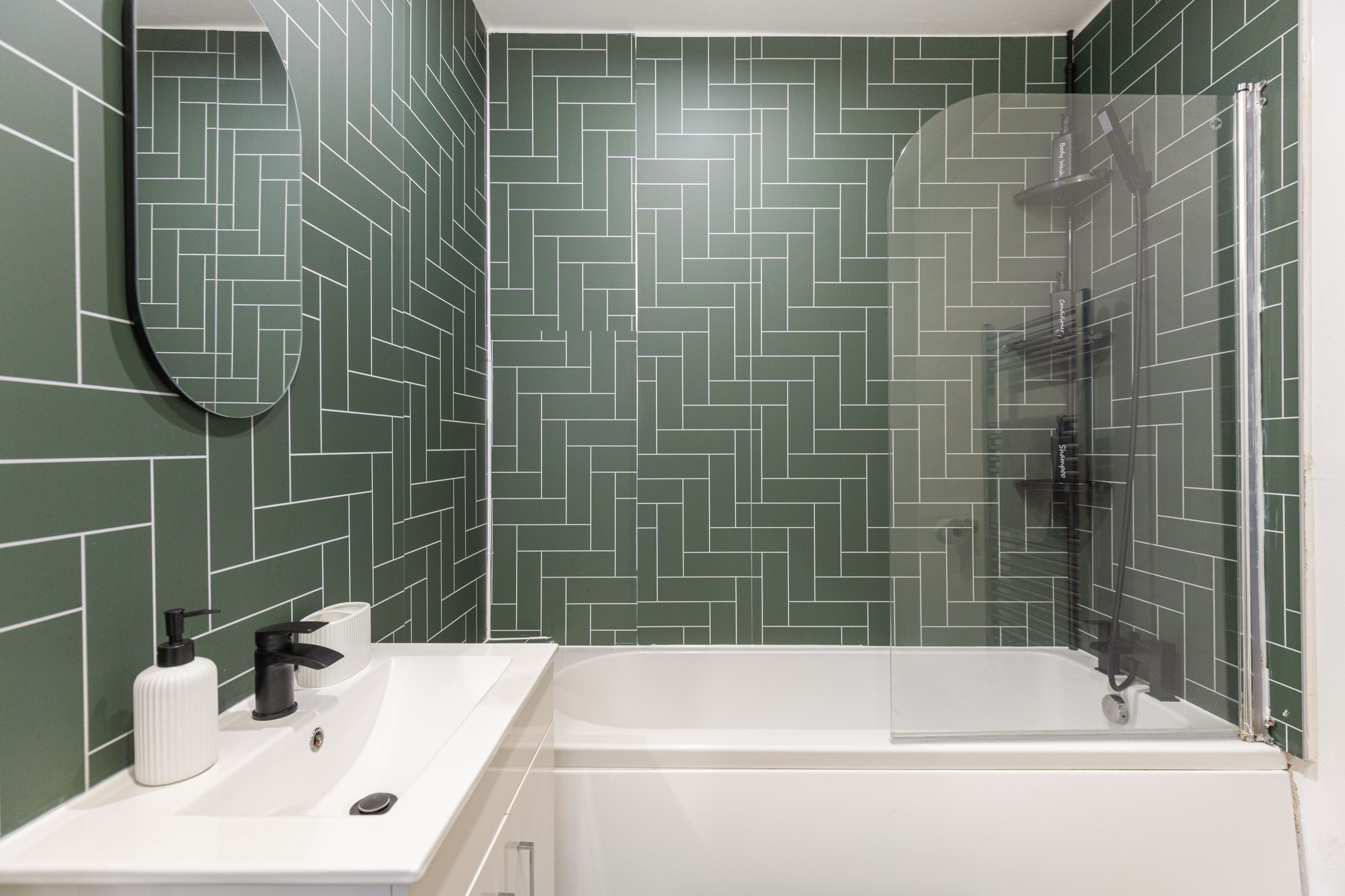 Une salle de bain avec un mur recouvert de carreaux disposés en motif chevrons, un lavabo blanc avec un miroir et une douche fermée par des parois en verre.