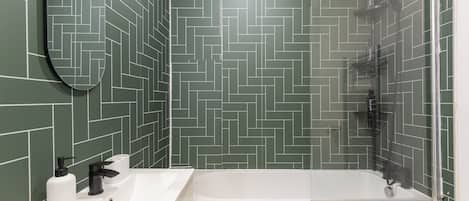 A bathroom with a herringbone-patterned tile wall, a white sink with a mirror, and a glass-enclosed shower.