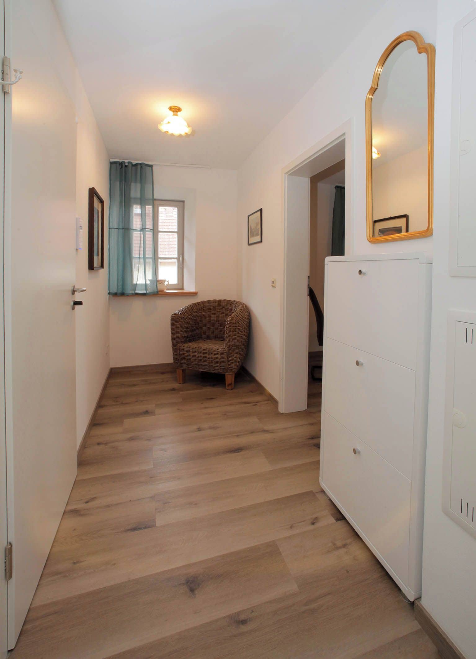 Un couloir avec un plancher en bois, une chaise en osier, une armoire blanche et un miroir au mur.