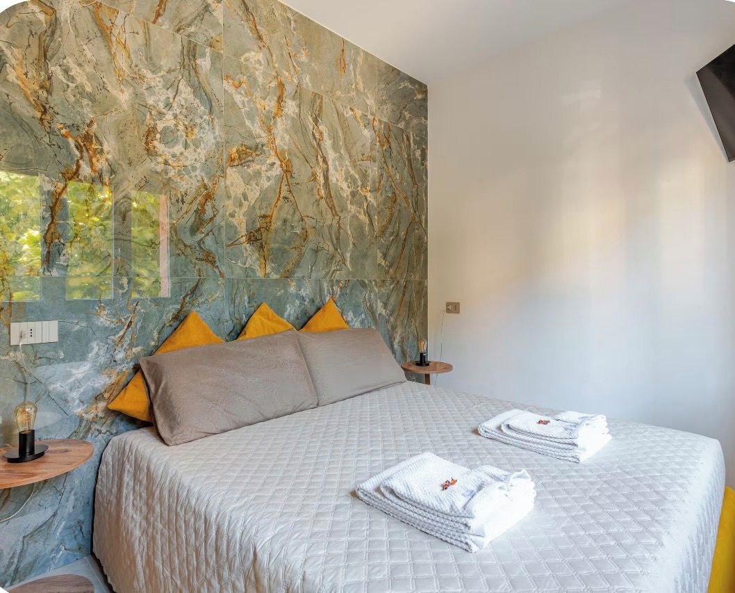 A hotel room with a marble wall, a bed with a quilted bedspread, two pillows, a bedside table with a lamp, and folded towels on the bed.