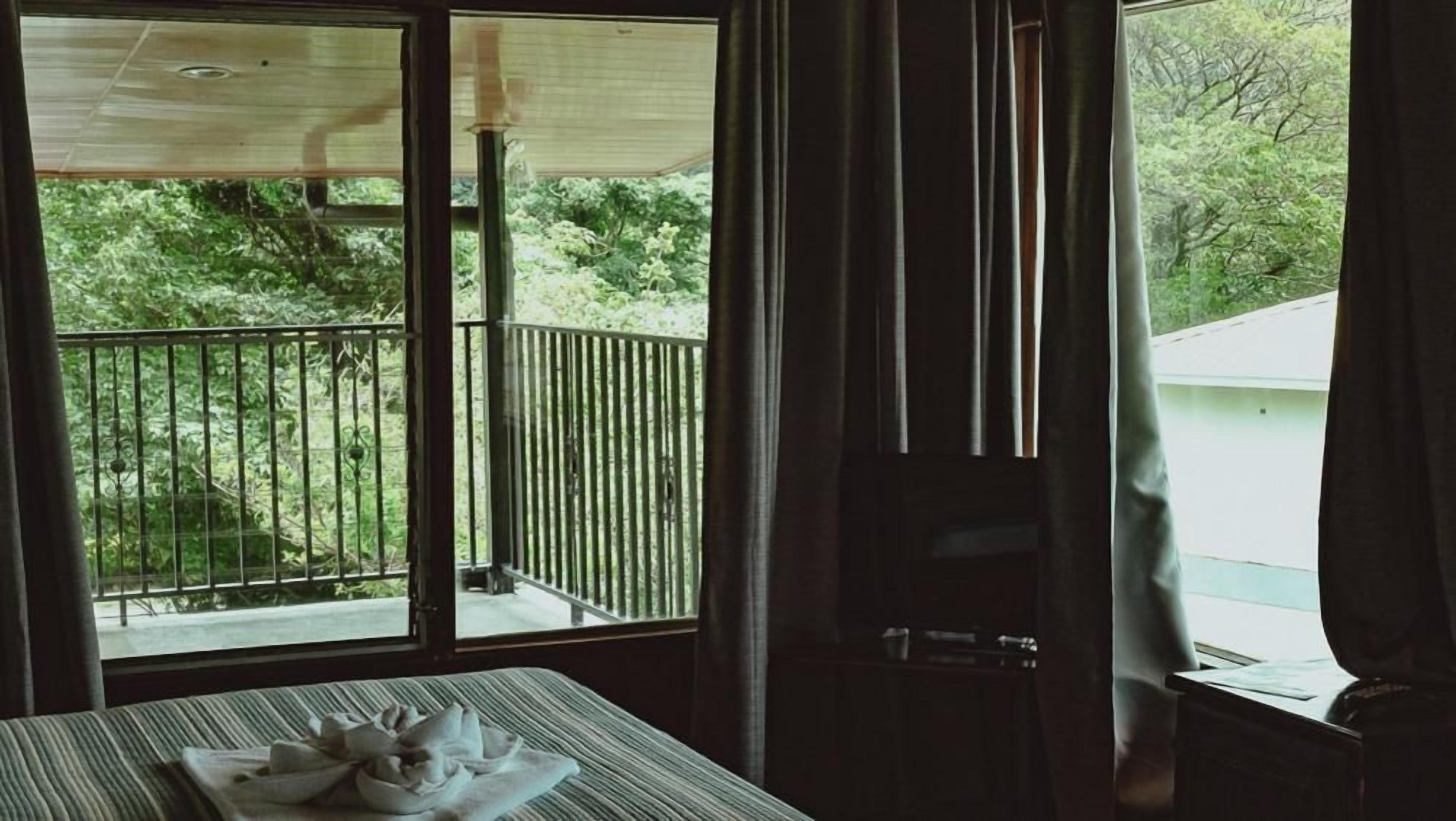 A room with a balcony, a bed, a table with towels, and a window with curtains.