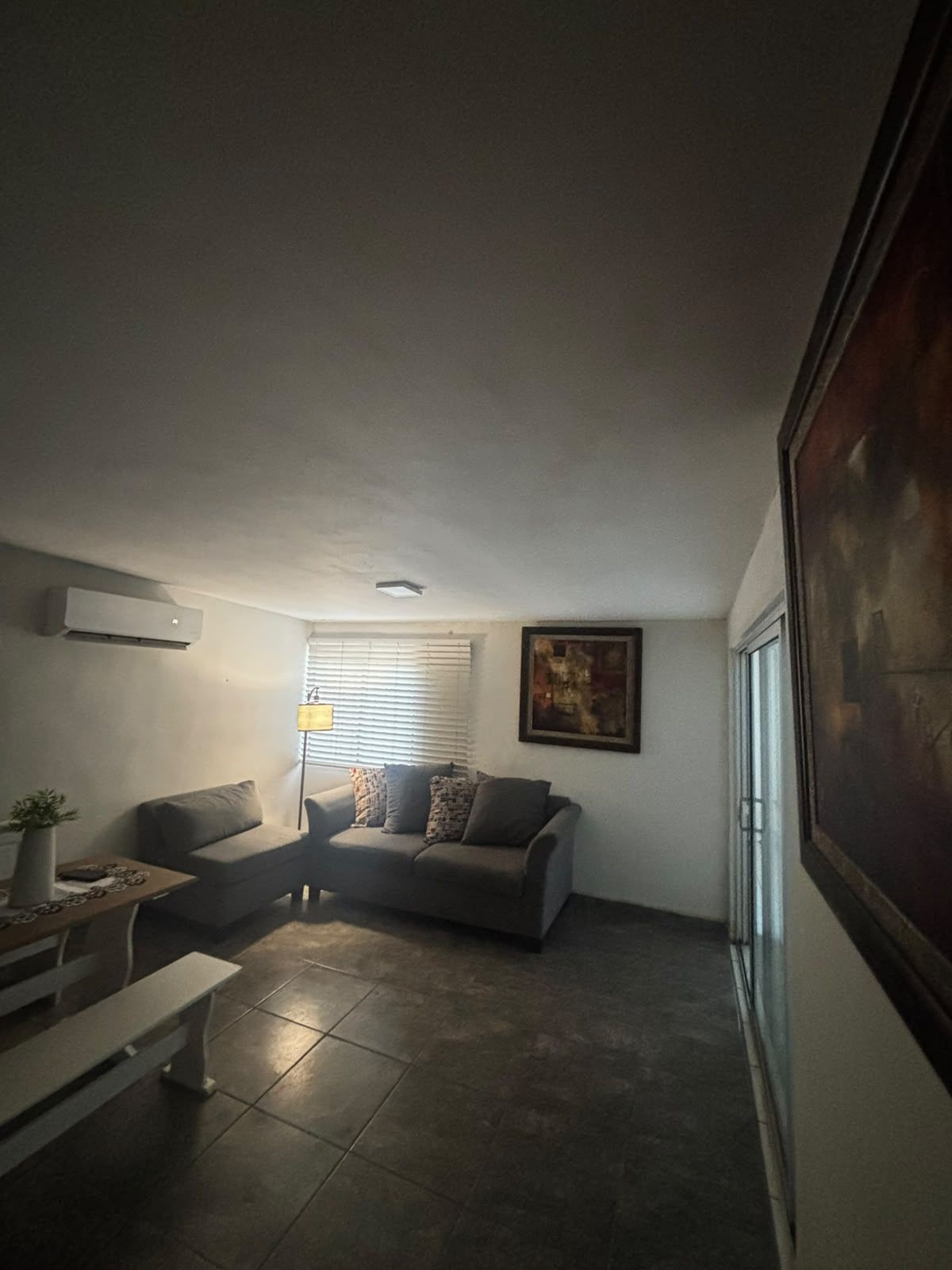 Una sala de estar con sofá, una mesa de centro y un banco. Hay un cuadro en la pared y una ventana con persianas.