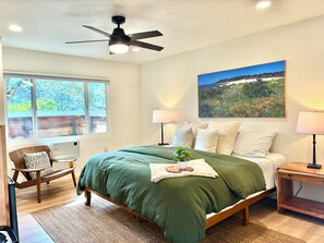 Ein Schlafzimmer mit einem Bett, Deckenventilator, einem Fenster mit Blick, einem Nachttisch mit Lampe und einem an der Wand hängenden Bild.