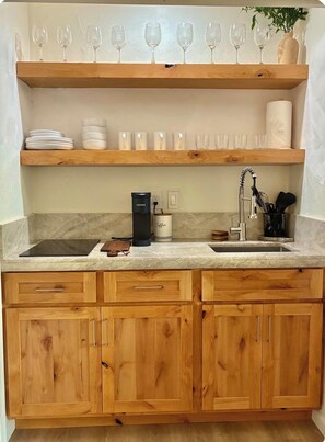 Una cocina con armarios de madera, encimera de mármol, una cafetera Keurig y una estantería con copas de vino y platos.