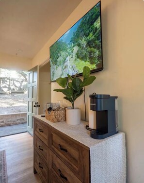 Ein Zimmer mit einem an der Wand befestigten Fernseher, einer Kaffeemaschine auf der Arbeitsfläche, einem Holzschrank und einer Topfpflanze.