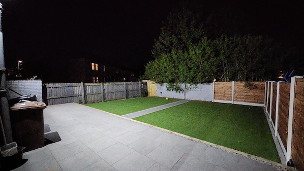 A backyard with a paved patio, a grassy lawn, and a wooden fence. - The Salford House (Salford)