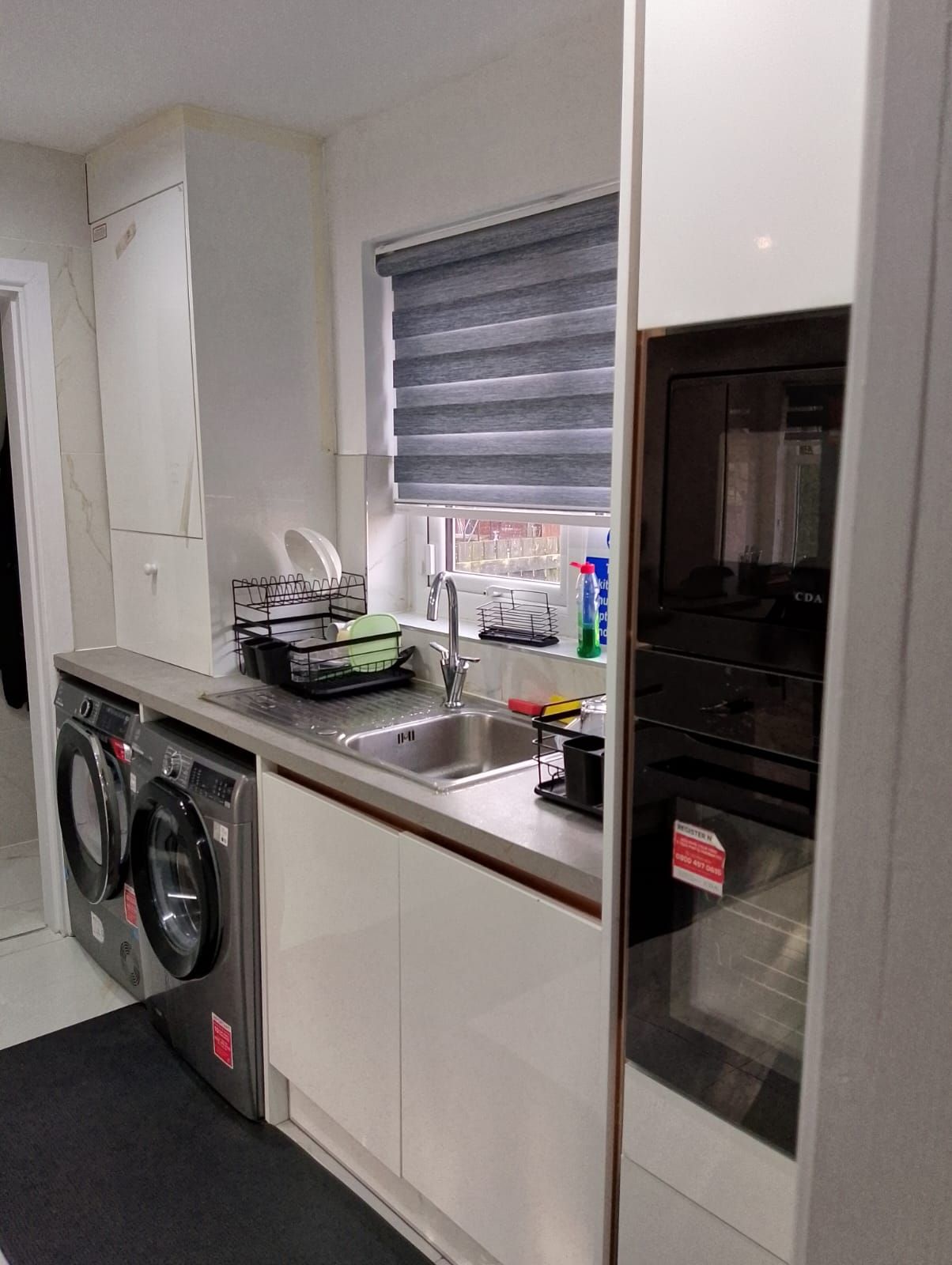 A compact kitchen with a built-in washing machine, a sink, and a window with blinds.