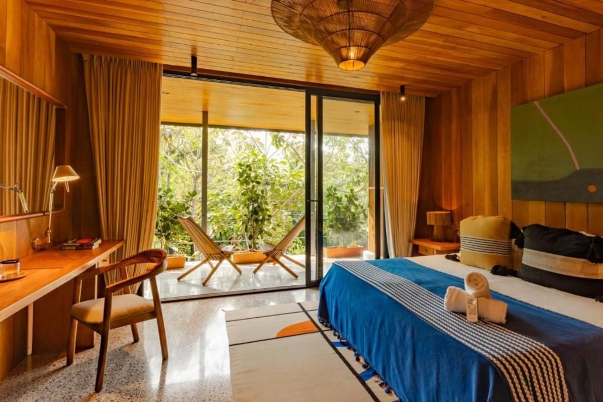 A hotel room with a bed, a desk, a chair, and a view of greenery through the window.