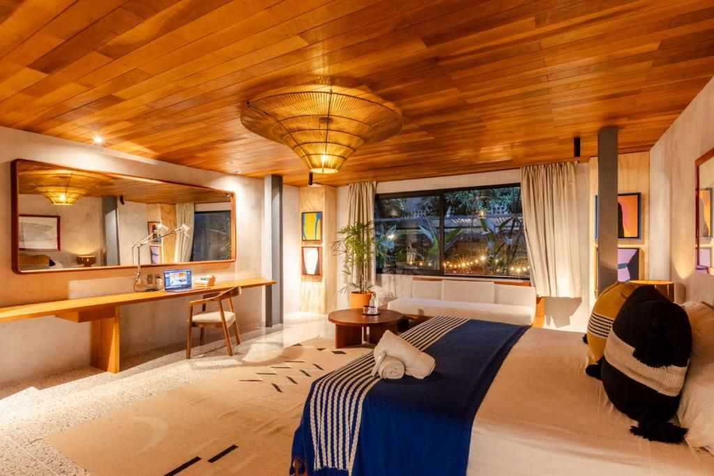 A hotel room with a wooden ceiling, a bed with a blue bedspread, a desk with a computer, a chair, and a window with curtains.
