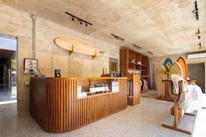 A reception area with a wooden counter, a surfboard, and a clothing rack with various items. - House of Somos, Santa Teresa (Santa Teresa)