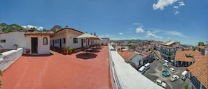 Eine Dachterrasse mit rotem Boden, ein kleines Gebäude mit einem Ziegeldach und Blick auf eine Stadt mit Gebäuden und blauem Himmel.