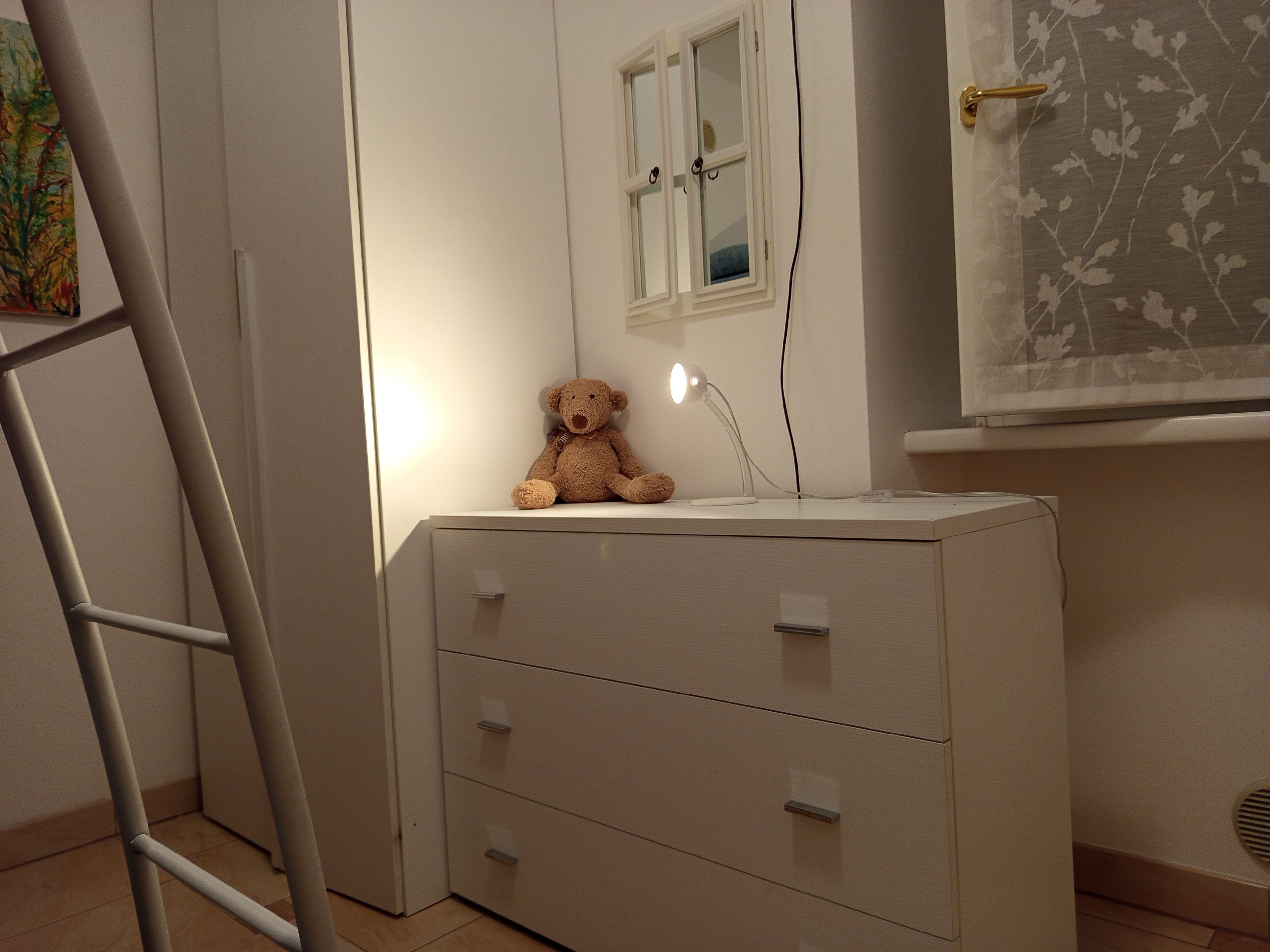 Une pièce avec une commode blanche, un ours en peluche posé dessus, une fenêtre avec des rideaux et une échelle appuyée contre le mur.