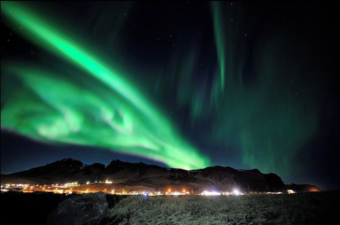 A night view of the aurora borealis over a mountainous landscape with a lit town below.