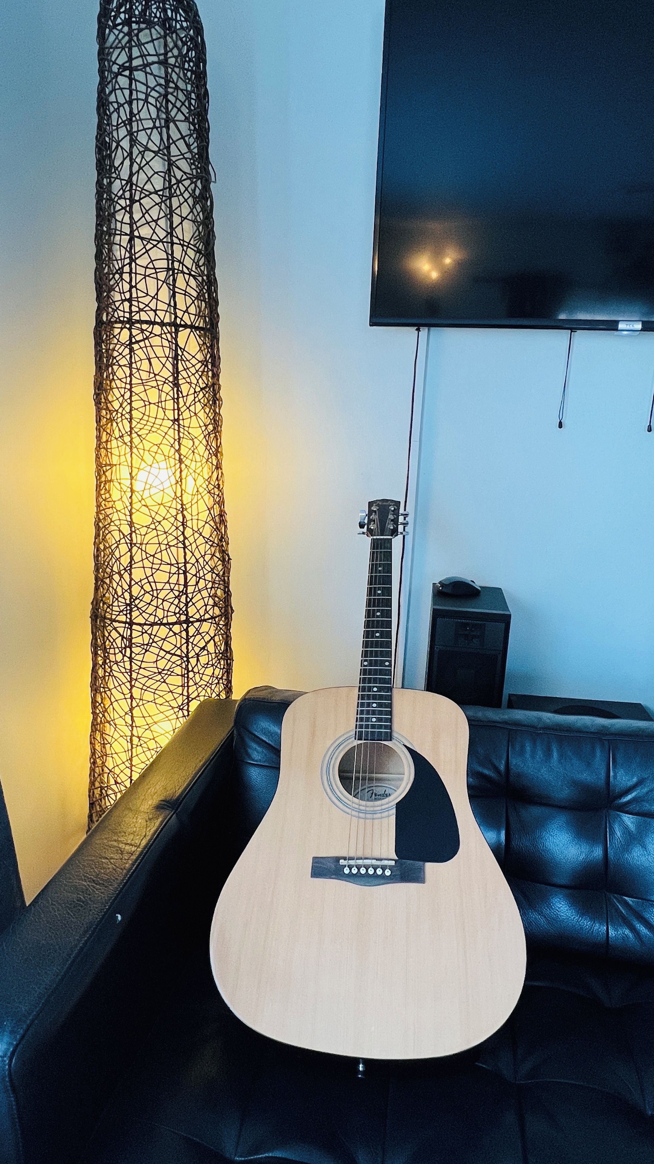 En guitar på en sort lædersofa med et vægmonteret fjernsyn og en høj, dekorativ lampe.