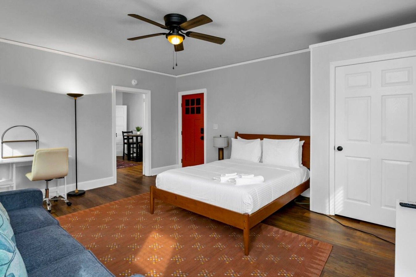 A hotel room with a bed, a desk, a chair, a ceiling fan, and a red door.