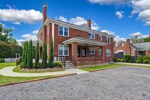 A two-story brick house with a front porch, a driveway, and a well-maintained lawn. - Walk to State St | 2 Beds + Queen Sleeper (West Columbia)