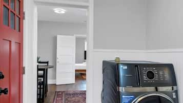 Una lavanderia con una lavatrice a carica frontale, una porta rossa e un corridoio che conduce a una camera da letto.