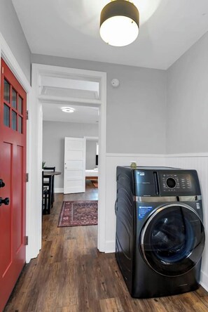 A laundry room with a front-loading washing machine, a red door, and a hallway leading to a bedroom. - Walk to State St | 2 Beds + Queen Sleeper (West Columbia)