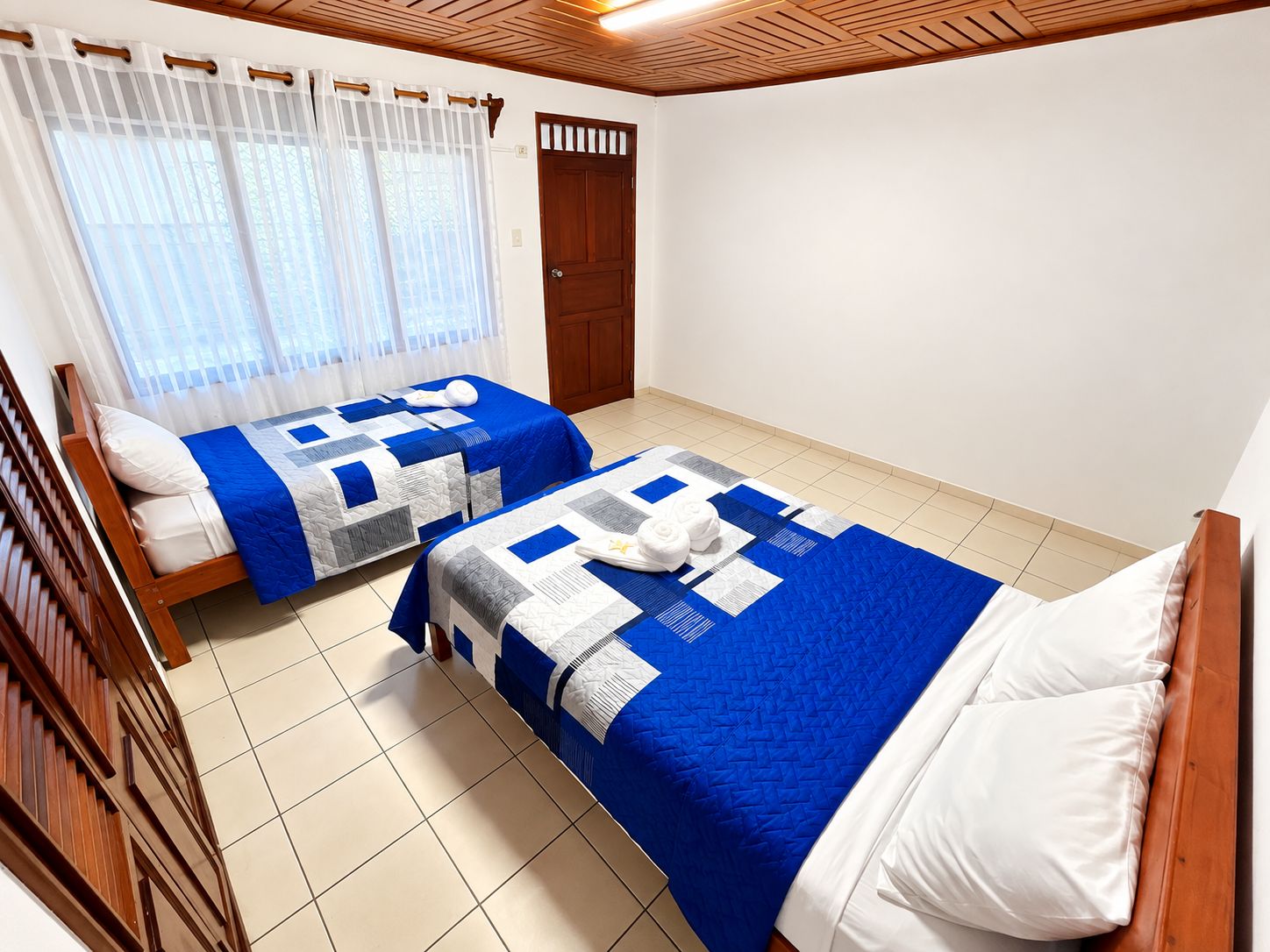 Une chambre d’hôtel avec deux lits, une tête de lit en bois, une fenêtre avec des rideaux légers et une porte avec un cadre en bois.