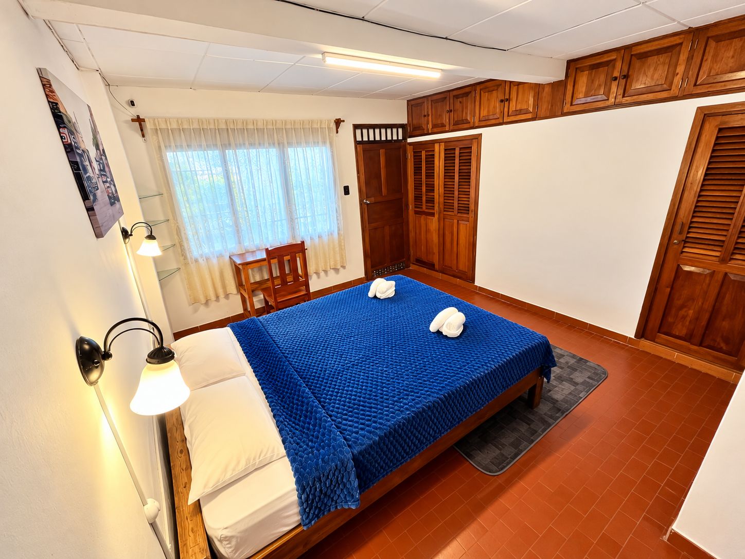 A bedroom with a bed, a desk, and a chair. There is a window with curtains, a door, and a wall-mounted light.