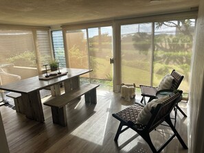 Una habitación amplia con un gran ventanal corredero que da a un jardín, una mesa de comedor de madera con bancos y una zona de estar con cojines y vistas.