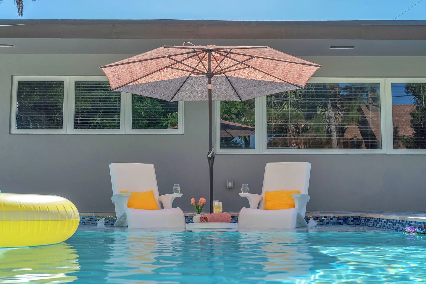 A swimming pool with two white lounge chairs, a pink umbrella, and a yellow inflatable float.