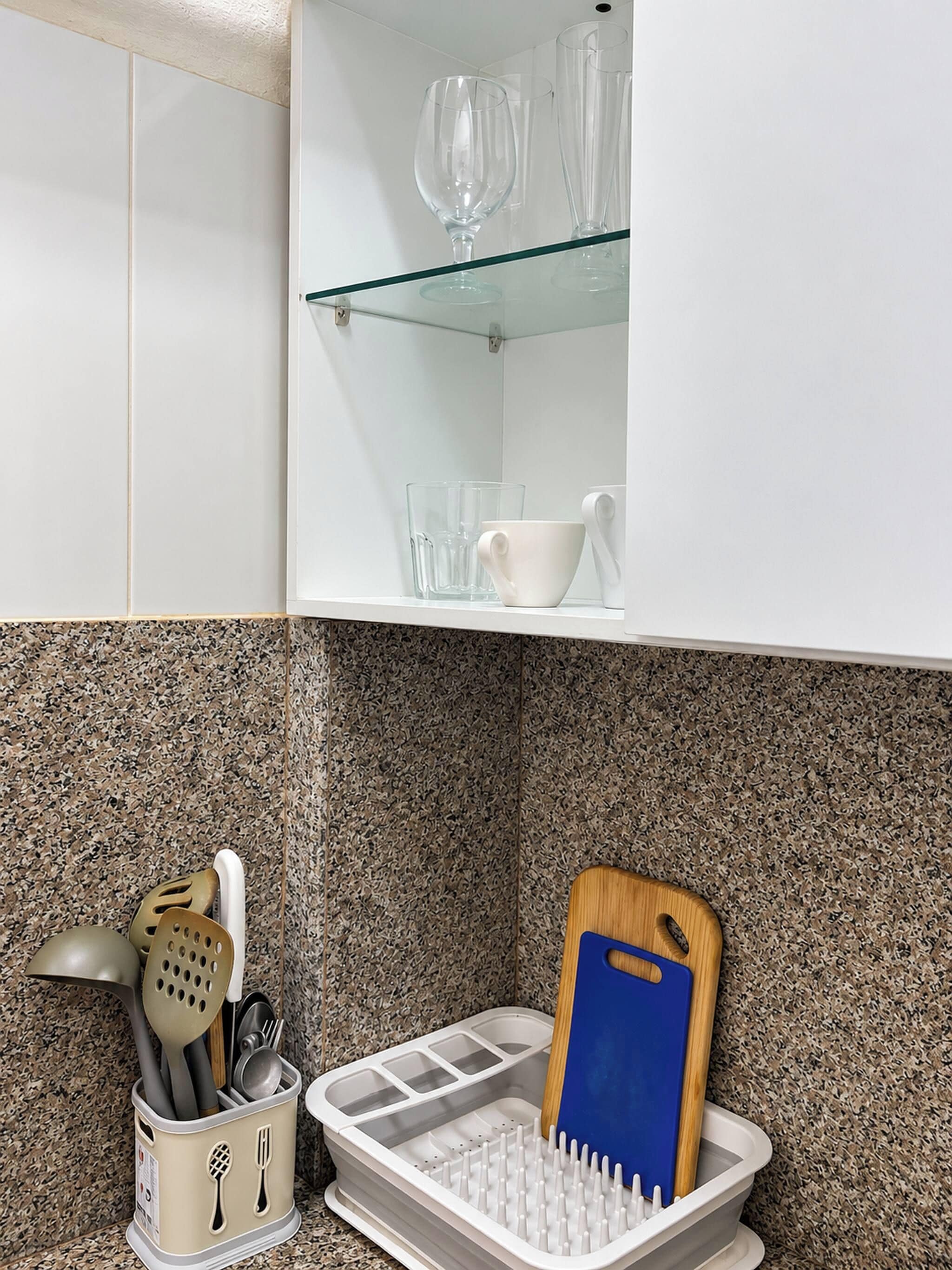 A kitchen with a white cabinet, a glass shelf, and a utensil holder with a blue cutting board.
